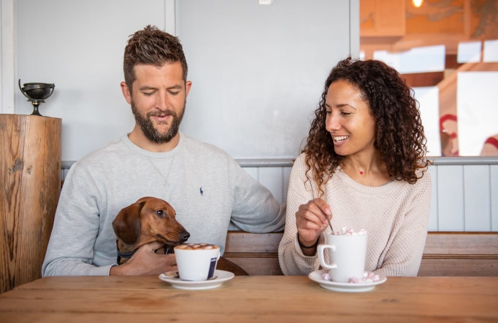 a man, woman and a dog sitting at a table with coffee cups in front of them